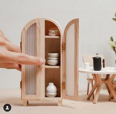 a hand is reaching into a dollhouse cupboard with dishes in it and a small table behind it