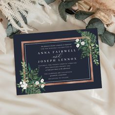 a wedding card with greenery and gold foil on it, next to some white flowers