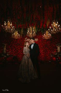 a man and woman standing next to each other in front of a red wall with chandeliers