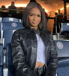 a woman wearing a black leather jacket standing in front of some blue seats with her hands on her hips