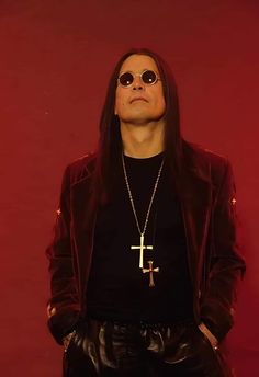 a man with long hair wearing sunglasses and a cross t - shirt standing in front of a red background