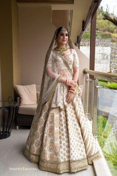 a woman in a white and gold bridal gown standing on a balcony looking at the camera