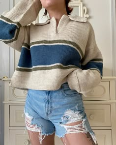 a woman wearing ripped shorts and a striped sweater poses for the camera with her hands on her head