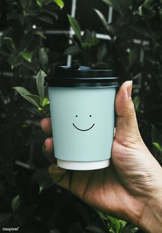 a person holding up a cup with a smiley face on the side and green leaves in the background