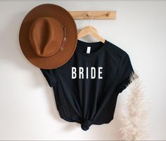 Black t-shirt on wooden hanger that reads BRIDE in white text. Between a brown hat and white plant. Future Mrs Shirt, Engagement Gifts For Bride, Bride Tee, Wedding Morning, Mrs Shirt, Bride Shirt, The Bachelorette, Custom Tee, Bridal Party Shirts