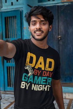 a man is pointing at the camera while wearing a t - shirt that says dad by day gamer by night