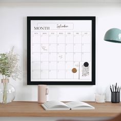 a desk with a calendar, pen and cup on it next to a plant in a vase