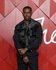 a man standing in front of a red wall wearing a fur collared jacket and black pants