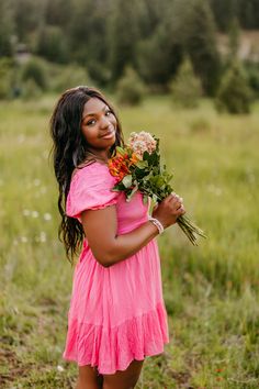 a woman in a pink dress holding flowers and smiling at the camera while standing in a field