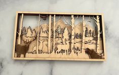 a wooden cutout of deer and trees with mountains in the background on a marble surface