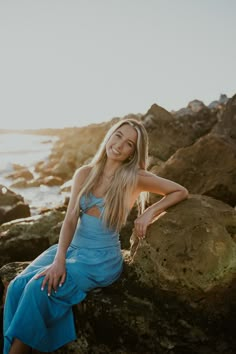 a woman in a blue dress sitting on some rocks by the ocean with her hand on her hip