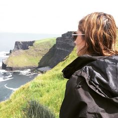 a woman standing on top of a lush green hillside next to the ocean with cliffs in the background