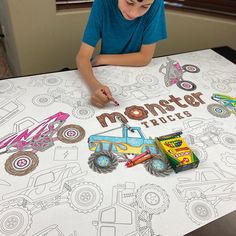 a young boy is sitting at a table with monster trucks on it and coloring paper