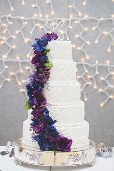 a white wedding cake with purple and blue flowers on the top tier is sitting on a silver platter