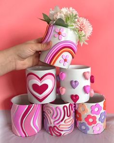 a hand is holding a flower pot with hearts and flowers on it, surrounded by other cups