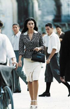 a woman walking down the street carrying a black purse and wearing white shoes with other people in the background