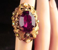 "Not for the shy, this HUGE brilliantly cut amethyst glass ring has a setting that measures a whopping 1 1/2\" and it raises up off your finger 1/2\". This is HUGE and stunning and is the prettiest purple you ever saw Around it are tiny rhinestones and pearls that really set off this magnificent setting. It is in great condition with rich patina and absolutely fit for a drama queen! Size 3 1/2 to a 5 since it is adjustable. Brass setting. See all SIX photos for more views and dimensions. 10-1-19 Vintage Oval Amethyst Ring For Wedding, Vintage Oval Amethyst Wedding Ring, Vintage Wedding Rings With Jewels, Vintage Amethyst Crystal Ring With Gemstone Detail, Vintage Amethyst Crystal Ring With Gemstone, Vintage Purple Amethyst Ring For Wedding, Vintage Purple Crystal Round Ring, Vintage Purple Crystal Ring, Vintage Purple Round Crystal Ring