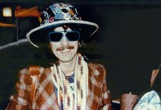 a man wearing sunglasses and a hat with beads around his neck, sitting in a chair