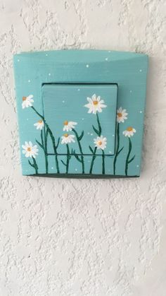 a painted wooden light switch cover with daisies on the inside and behind it is a blue frame