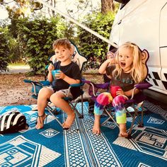 two young children sitting in lawn chairs on a blue and white rug next to a van