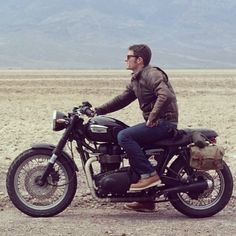 a man riding on the back of a motorcycle across a desert field with mountains in the background