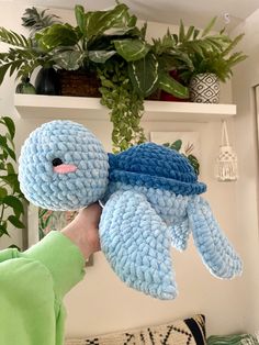 a person holding up a blue crocheted stuffed animal in front of a potted plant