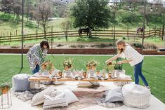 two women standing around a table with flowers and plants on it in the grass near a fence