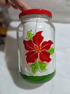 a hand painted glass jar with red and green flowers on the lid is being held by a person