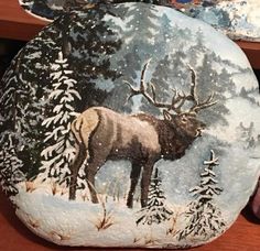 a painted rock with an image of a moose and pine trees in the snow on it