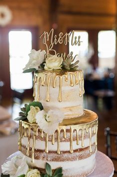 a wedding cake with white flowers and gold icing on it's top is sitting on a table