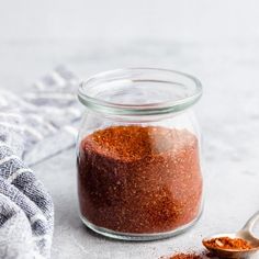 a glass jar filled with red chili seasoning next to a spoon and cloth on the table