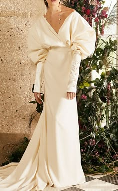 a woman in a white dress standing next to a wall with flowers and greenery