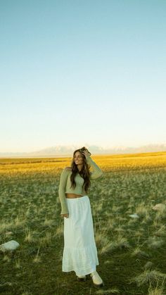 a woman standing in a field wearing a white skirt and cropped top with her hands on her head