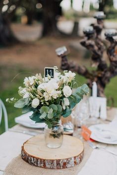 the table is set with white flowers and greenery in a vase on top of a wooden slice