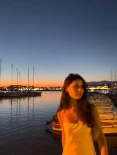 a woman in a yellow dress is standing by the water at night with boats and buildings in the background