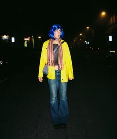 a woman with blue hair wearing a yellow jacket and scarf standing in the street at night