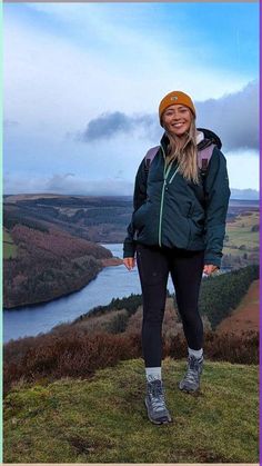 a woman standing on top of a grass covered hill next to a lake and mountains