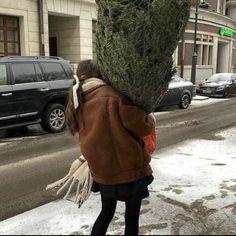 a woman walking down the street carrying a christmas tree on her head and wearing a brown jacket