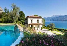 an outdoor swimming pool surrounded by flowers and greenery with a house in the background