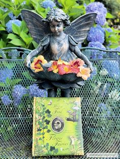 a statue of a fairy sitting on top of a book in front of some flowers