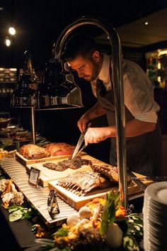 a man in an apron cuts up food at a buffet table with other foods on it