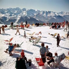 a group of people sitting on top of a snow covered slope with mountains in the background