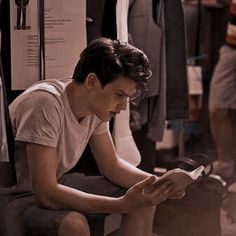 a young man is reading a book while sitting on a chair in front of some clothes