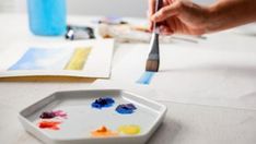 someone is painting on a plate with paintbrushes and watercolors in front of them