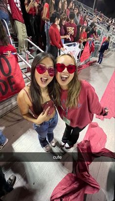 two girls wearing red sunglasses at a football game with fans in the stands behind them