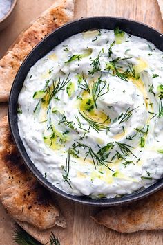 a bowl filled with white dip surrounded by pita bread on a wooden table top