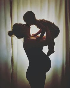 a woman holding a baby in her arms while standing next to a curtain with the light coming through it