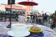 a cup of coffee and a croissant on a table with an umbrella in the background