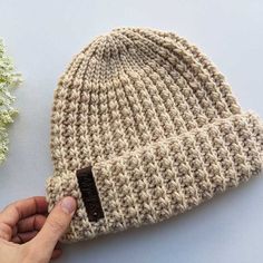 a hand is holding onto a knitted beanie with a tag on it, next to some flowers