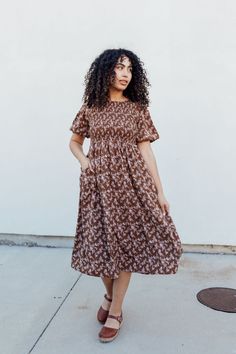 Stay stylish with the Emsley Midi dress! Featuring a charming brown color with delicate white florals, this dress is designed with a smocked bodice and flared bubble sleeves for a fun and flattering look. Plus, it has pockets for added convenience. Look effortlessly chic in this must-have dress! The Emsley Midi is lined. Fits true to size. Model is wearing a small and is 5'10". Plus model is wearing a 2X and is 5'6". Size Chart Length Bust Waist S 46" 30" 28" M 46" 32" 30" L 47" 34" 32" XL 48" 3 Brown Knee-length Ruffled Midi Dress, Brown Ruffled Knee-length Midi Dress, Brown Modest Midi Dress For Spring, Modest Brown Midi Dress For Spring, Fall Brown Midi Dress With Puff Sleeves, Brown Midi Length Dress With Smocked Back, Brown Ruffled Dress For Day Out, Brown Short Sleeve Dress With Pockets, Brown Short Sleeve Dress For Garden Party